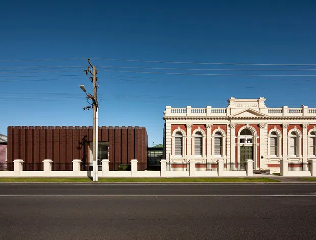 Street-facing view of a historic ornate neoclassical-style building with a cream and muted red facade, tall arched windows, decorative pilasters, balustrades, and a parapet. A modern dark vertical-panel volume sits immediately beside it, creating a contrasting heritage and contemporary frontage. The scene is viewed from across a broad paved road with overhead utility wires and a low front fence.