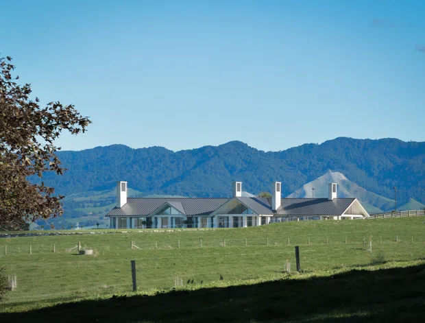 A long, low rural property set behind expansive fenced pasture, with multiple connected gabled roof forms, pale exterior walls, dark grey metal roofing, and several prominent chimneys. Forested mountains rise behind the building under a clear blue sky, creating a distinctly alpine or rural landscape setting.