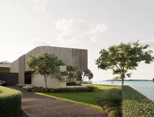 A contemporary two-storey waterfront residence with a strongly rectilinear form, vertical timber-like facade screening, pale masonry walls, and dark recessed glazing. The home is approached by a curved paved driveway and surrounded by manicured hedges, lawn, sculptural shrubs, and mature trees, with a calm body of water directly beyond the property.