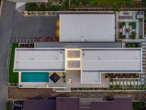 Aerial overhead view of a contemporary residential property, showing several pale metal-roofed wings arranged around landscaped courtyards and paved paths. A rectangular swimming pool with adjacent umbrella is visible on the left, with structured garden beds, outdoor lighting, and surrounding driveways and neighbouring roofs.