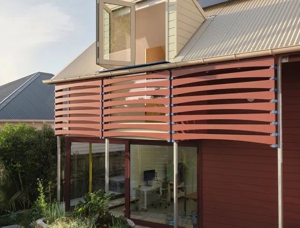Exterior view of a contemporary multi-level home with a steep corrugated metal roof, dark solar panels, red-brown weatherboard cladding, and a distinctive upper-level dormer window. A curved horizontal timber privacy screen projects over glazed ground-floor living spaces, opening onto a timber deck and landscaped garden.