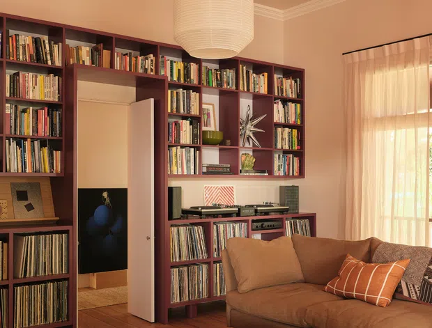 Warm, richly coloured living room featuring extensive dark burgundy built-in bookshelves filled with books and vinyl records, a turntable and stereo equipment, a large brown sectional sofa, leather ottomans, timber flooring, and a central oversized paper pendant light. Decorative ceiling mouldings and coffered detailing frame the room, while sheer curtains filter daylight through a tall window or glazed door.