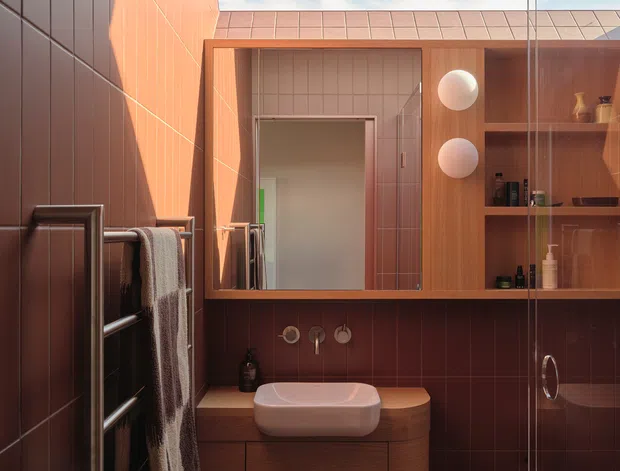 Compact contemporary bathroom with warm terracotta wall and floor tiles, a timber floating vanity with a white vessel basin, wall-mounted fixtures, open timber shelving, and a glass shower enclosure. A skylight or glazed roof opening brings in daylight, while a heated towel rail and partially visible toilet complete the space.