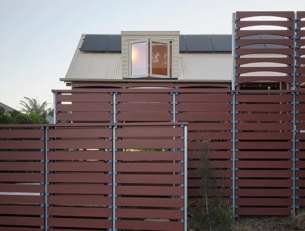 Exterior view of a residential property screened by a tall horizontal-slat fence. The fence uses dark reddish-brown boards with visible blue-grey metal posts and framing, including a taller curved-top privacy screen at right. Behind it is a light corrugated pitched roof with a small dormer window, while low landscaping and a paved walkway occupy the foreground.