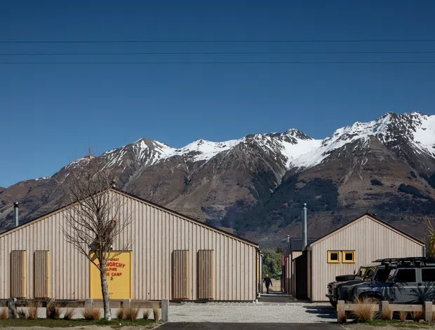 Exterior view of a group of simple gable-roofed, single-storey buildings with vertically ribbed light-coloured cladding, set against a dramatic snow-capped mountain range. The foreground includes a gravel driveway, sparse landscaping, utility elements, and parked vehicles. The image is predominantly an architectural exterior scene.