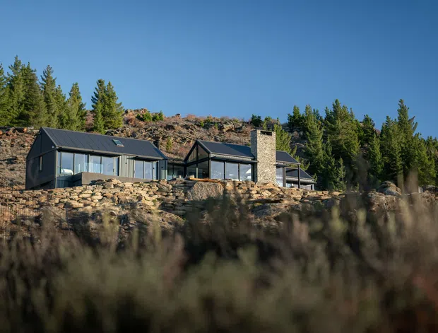 A low-slung contemporary house set among exposed rocky terrain and evergreen trees. The building has dark metal pitched roofs, extensive glazing, dark cladding, and a prominent tall stone chimney. It is photographed from across a blurred natural foreground, with a rugged wooded hillside behind.