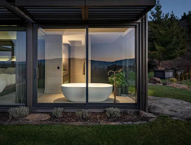 Exterior view of a contemporary glass-walled bathroom pavilion at dusk, with a freestanding white bathtub prominently visible behind floor-to-ceiling glazing. The reflective glazing also reveals a bedroom with a bed on the left. A dark steel-framed pergola or roof canopy projects over the facade, while landscaped planting, lawn and a stone path surround the structure. Mountainous terrain is visible in the reflections and background.