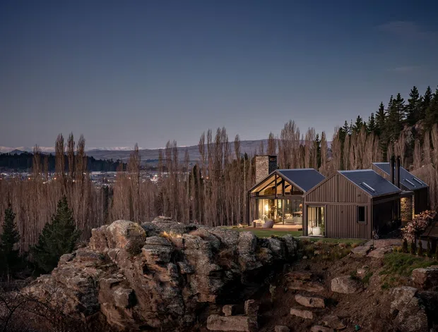 A modern multi-building house set among rocky outcrops and tall trees on a wooded, elevated site. The buildings have dark vertical cladding, steeply pitched gabled roofs, and extensive glazed sections, with distant hills visible beyond. The scene is photographed at dusk, creating a dramatic, subdued atmosphere.