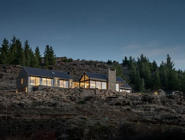 A modern single-storey house set into a rugged rocky, forested hillside at dusk. The building has dark pitched roofs, extensive warm-lit glazing, timber-framed gabled sections, and a prominent stone-clad chimney or masonry feature. The surrounding exposed rock and coniferous trees create a secluded mountain or bush setting.