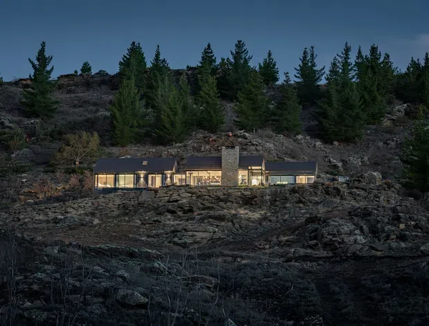 A low, modern single-storey house embedded in a rugged rocky hillside, photographed at dusk. The elongated building has dark pitched roof planes, extensive warm-lit glazing, and a prominent central stone or concrete chimney. Conifer trees and exposed dark rock surround the home, creating a secluded alpine or forested setting.