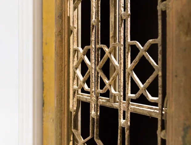 Close-up architectural detail of an aged, ornate metal security grille or window gate with geometric diamond and X-pattern bars, set within a weathered timber or painted frame. The distressed surface shows layered cream, ochre, and brown tones, with a dark opening behind it and a narrow white trim at the left edge.