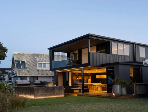 Exterior view of a contemporary two-storey home at dusk, featuring a dark-clad upper level, broad glazed openings, a covered balcony, and a warm, open-plan living and kitchen area visible on the ground floor. The house sits in a landscaped suburban setting beside an older corrugated-roof dwelling, with a low rendered retaining wall and lawn in the foreground.