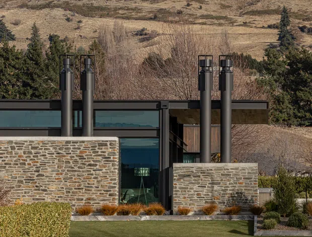A modern low-profile house set against a dry, hilly landscape. The facade combines dark metal framing and roof elements, extensive glazing, and prominent natural stone walls, with multiple exposed cylindrical chimney flues rising above the roofline. Structured landscaping and lawn occupy the foreground.