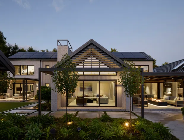 A dusk view of a large contemporary residential house arranged around a landscaped courtyard. The central gabled pavilion has a dramatic glazed facade with horizontal timber screening, flanked by low-pitched roof wings and covered outdoor living areas. Dark metal roofing, pale rendered or masonry walls, extensive glazing, structured planting, and warm interior and landscape lighting create a refined indoor-outdoor setting.