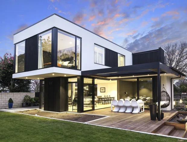 Contemporary two-storey residence photographed from the rear garden, featuring crisp white upper-level volumes, dark vertical cladding, extensive floor-to-ceiling glazing, a covered outdoor dining terrace, and timber decking with integrated planters. The landscaped backyard includes a lawn, paved areas, and a low masonry boundary wall.