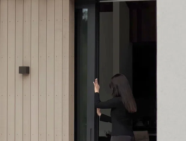 Close-up exterior view of a contemporary building entrance with pale vertical timber cladding, dark aluminium-framed sliding glass doors, and a projecting dark metal canopy with exposed structural beams. A person is opening the door, with a dim interior and furniture faintly visible through the glazing. A sculptural pale outdoor stool or table sits in the foreground.