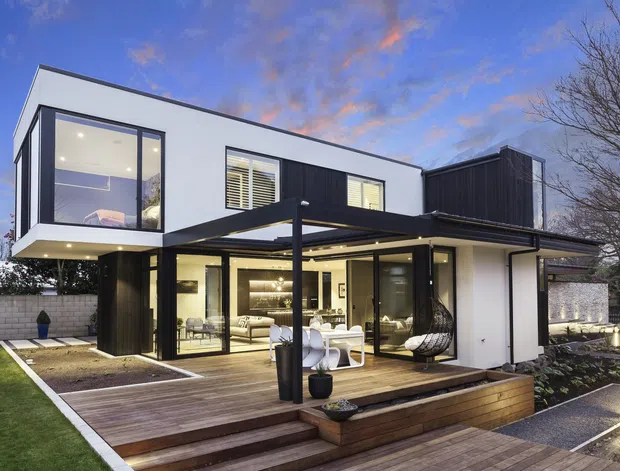 Contemporary two-storey home exterior at dusk, featuring crisp white rendered walls, dark vertical cladding, large glazed openings, and a projecting upper-level box. A covered outdoor entertaining area opens onto a timber deck with built-in steps and planters, while the illuminated living spaces are visible through the glazing.