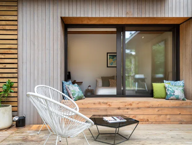 A warm contemporary timber-clad exterior seating nook with a large dark-framed sliding window opening onto an interior living room. The facade combines vertical weathered timber boards with horizontal timber screening and a built-in timber window seat, furnished with a white sculptural wire lounge chair, patterned cushions, and a small black coffee table. Potted plants and natural timber tones create an inviting indoor-outdoor connection.