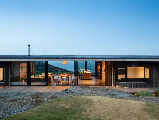 Single-storey contemporary coastal house viewed from the landscaped front, with a long low flat roof, dark vertical cladding, extensive floor-to-ceiling glazing, and warm timber interiors visible through the glass. The home opens toward a rocky coastal landscape and ocean horizon, with an outdoor dining area and central living space apparent behind the glazing.