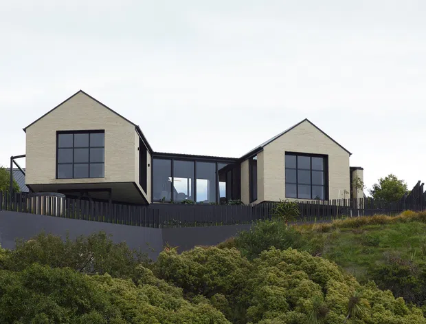 Exterior view of a contemporary two-wing residential house on a sloping, vegetated site. The home features two steep gabled volumes clad in pale masonry or textured panels, connected by a darker glazed central section, with large black-framed windows, a dark fence, and elevated foundations.