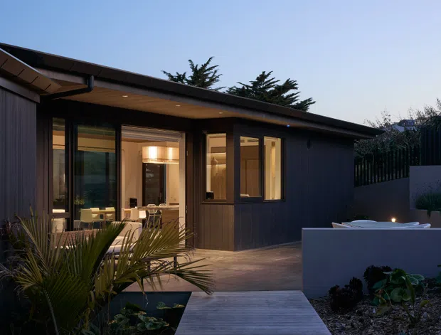 A dusk exterior view of a low-profile contemporary pavilion or extension with dark vertical cladding, broad roof overhangs, warm recessed lighting, and large glazed openings. The glazing reveals a warmly lit interior dining or living area. A timber walkway crosses a landscaped garden toward the building, with a raised pale rendered outdoor feature or planter in the foreground.