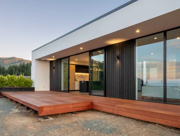 A contemporary single-storey house with a flat roof, crisp white rendered walls, dark vertical timber cladding and extensive black-framed floor-to-ceiling glazing. A warm timber deck wraps the frontage, with an illuminated interior visible through the glass, including a bedroom and kitchen or living area. The home sits in a landscaped setting with hills visible in the distance.