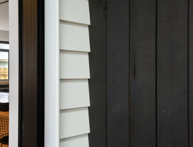 Close-up architectural view of a dark vertical timber-clad exterior wall beside light horizontal weatherboard cladding and a dark-framed opening. Through the opening, part of a modern kitchen is visible, including a stone island, sink, cabinetry and wire dining chair. The image emphasizes contrasting facade materials and the connection between exterior cladding and interior space.