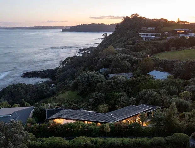 Elevated dusk view across a rugged coastal headland, with the ocean and rocky shoreline on the left and several homes dispersed among dense native vegetation and manicured lawns. The foreground features a low, sprawling contemporary house with dark pitched roofing, extensive glazing, and warm interior lighting, set into the sloping landscape.