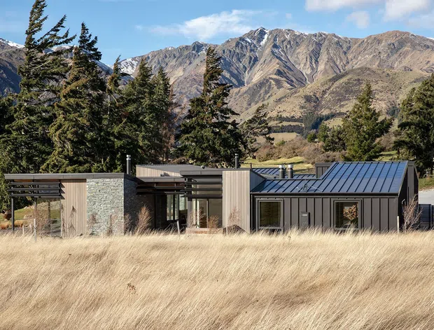 A low-profile contemporary mountain house set among tall conifers and dry golden grasses, with dramatic rugged alpine mountains behind it. The building combines dark vertical metal cladding, pale timber panels, grey stone walls, broad glazing, and a low-pitched standing-seam metal roof. Its horizontal composition and partially open timber screens blend into the rural landscape.