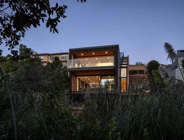 Dusk exterior view of a contemporary multi-level home, partially screened by dense ornamental grasses and vegetation. The house features extensive warm-lit glazing, projecting upper-level balcony or living space, dark structural framing, timber-toned lower cladding, and a flat roof. Nearby residential buildings and coastal-looking planting are visible in the background.