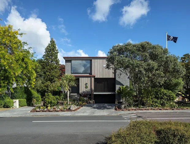 Street-facing view of a contemporary multi-level residence set behind landscaped planting. The house has a strong rectilinear form, a low-pitched or flat roofline, dark vertical cladding, large upper-level glazing, and a recessed dark garage or entry opening. Mature trees and garden beds frame the frontage, with a paved driveway connecting to the street. A flag is visible above the property.
