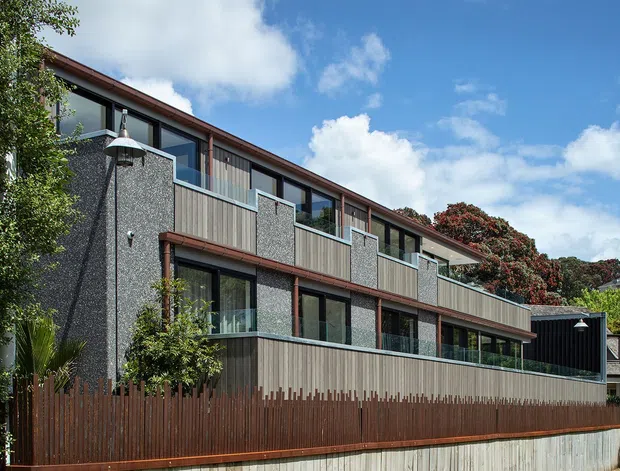 Exterior view of a contemporary multi-storey residential building on a sloped urban or suburban site. The facade combines dark grey textured wall panels, vertical timber cladding and extensive dark-framed glazing, with projecting balconies and a long low-pitched roofline. A timber privacy fence and landscaped planting occupy the foreground.