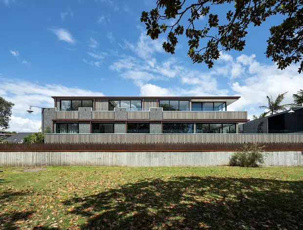 Exterior view of a contemporary multi-storey residence with a broad flat roof, extensive floor-to-ceiling glazing, projecting upper-level balconies, and a textured grey masonry or concrete facade. The property is seen behind a timber privacy fence and landscaped lawn, with mature trees framing the view.