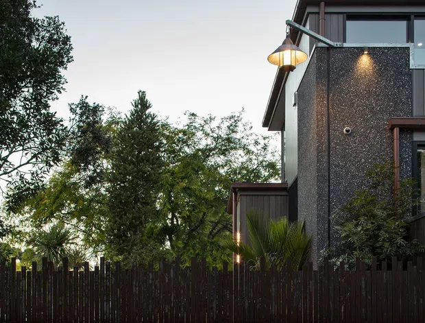 Exterior view of a contemporary multi-level home at dusk, partially screened by mature landscaping and a dark vertical timber fence. The facade combines dark textured masonry or render with vertical dark cladding, large glazed openings, projecting roof forms, and warm exterior lighting.