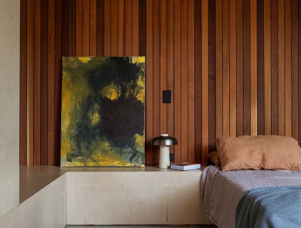 A warm, moody bedroom with a full-height vertical timber slat feature wall, pale built-in platform cabinetry and bedside ledge, polished grey concrete flooring, and a partially visible bed dressed in muted taupe and grey textiles. An abstract artwork and compact table lamp sit on the ledge, creating a restrained, contemporary interior.