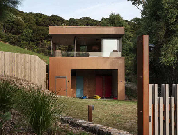 A compact contemporary two-storey house set into a heavily vegetated, sloping site. The facade is clad in warm brown timber-toned panels, with a broad projecting flat roof, large upper-level glazing, and an open ground-level garage or storage bay containing colourful objects. A timber privacy fence, stone-edged landscaping, and a tall matching entry or gate element frame the foreground.