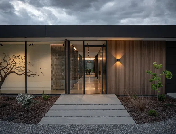 A dramatic contemporary single-storey building entrance viewed from a landscaped forecourt. The facade combines dark horizontal cladding with extensive floor-to-ceiling glazing, a warm timber-look vertical feature wall, and a long illuminated corridor visible through the entry. A concrete stepping-stone path crosses a gravel and planted garden, while a sculptural tree mural and interior artwork add visual interest. Heavy storm clouds create a moody architectural setting.