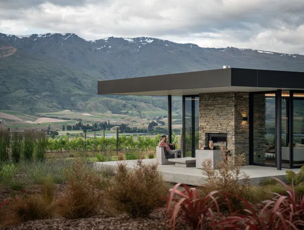 A contemporary single-storey pavilion-style home set among landscaped planting and a vineyard, with dramatic mountain ranges in the background. The building features a broad flat roof, extensive floor-to-ceiling glazing, slender dark frames, and a central stacked-stone fireplace wall. An outdoor terrace extends from the living area.