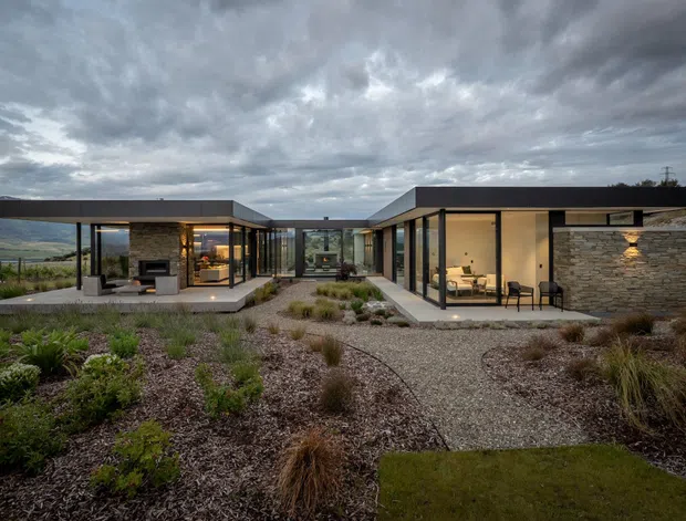 A low-profile single-storey modern residence arranged around landscaped outdoor areas, photographed at dusk beneath a dramatic cloudy sky. The building features flat roof planes, extensive floor-to-ceiling glazing, dark slender frames, warm interior lighting, and a prominent natural stacked-stone wall. Gravel paths, native-style planting, ornamental grasses, and clipped lawn connect the pavilion-like wings. A living area with a fireplace and furnishings is visible through the glazing.