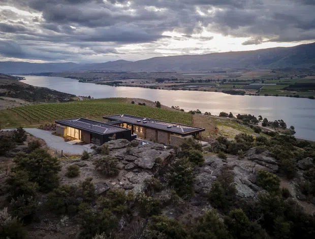 A low-profile contemporary house with dark flat roofs and extensive glazing sits among rocky scrub on a sloping hillside above a broad lake. The remote landscape includes vineyards or cultivated fields, rugged mountains, and dramatic overcast evening light.