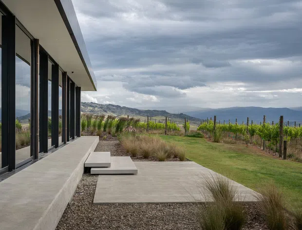A low, modern pavilion-like building extends along the left side of the image, with extensive dark-framed floor-to-ceiling glazing beneath a projecting flat roof. A broad concrete terrace and stepping slabs lead through landscaped ornamental grasses toward a lawn and vineyard, with layered hills and a cloudy sky beyond. The composition emphasizes the building’s strong horizontal lines, indoor-outdoor connection, and rural landscape setting.