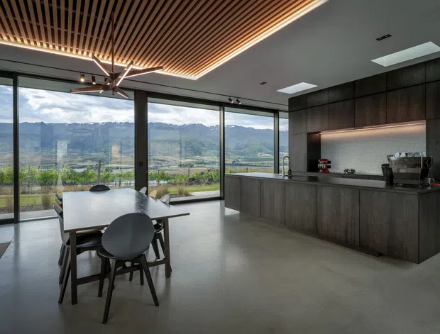 A contemporary open-plan kitchen and dining area with expansive floor-to-ceiling glazing overlooking a mountainous landscape. The space features dark wood kitchen cabinetry and island joinery, a smooth concrete floor, a large white dining table with dark timber chairs, and a striking slatted timber ceiling with integrated linear lighting. A skylight and minimalist track lighting add illumination, while the broad glazing creates a strong indoor-outdoor connection.