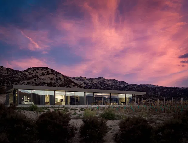 A low, elongated modern house set against rugged mountainous terrain at dusk. The building has extensive floor-to-ceiling glazing, a dark slender roofline, and a transparent pavilion-like form. A cultivated garden or vineyard lies in the foreground, while a dramatic pink and purple sunset sky dominates the background.