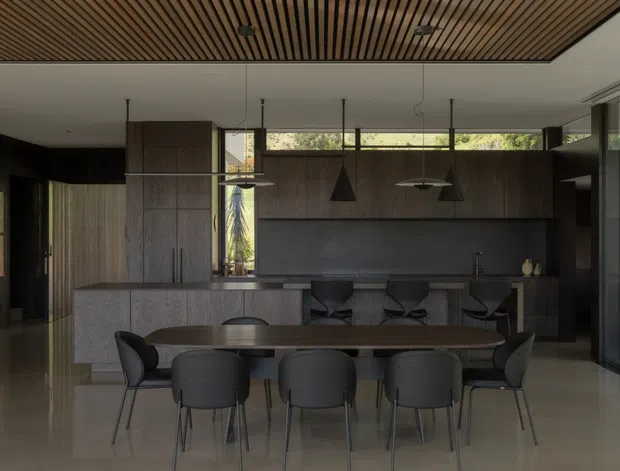 A dark, refined open-plan dining and kitchen interior. A long timber dining table with six upholstered chairs sits in the foreground, while a minimalist kitchen with a large island, integrated dark cabinetry, stone surfaces and bar seating occupies the background. Floor-to-ceiling glazing brings in filtered greenery, and a slatted timber ceiling adds warmth and texture. Pendant lights hang above both the island and dining area.