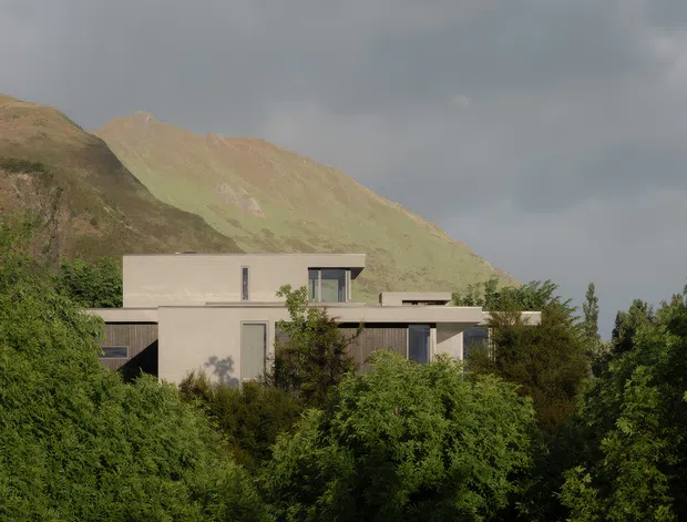 A modern flat-roofed, multi-level house partially concealed by dense greenery, set against a dramatic mountain landscape beneath an overcast sky. The building has simple rectilinear volumes, pale rendered walls, dark-framed windows, and restrained contemporary detailing.