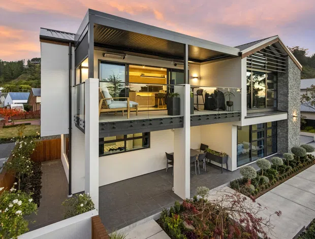 Contemporary multi-level residential home viewed from the exterior at dusk. The facade combines white rendered walls, dark metal framing, extensive glazing, a projecting balcony with outdoor seating, and a prominent pitched gable volume clad in textured grey stone. Landscaped gardens, paved paths, and neighbouring homes surround the property.