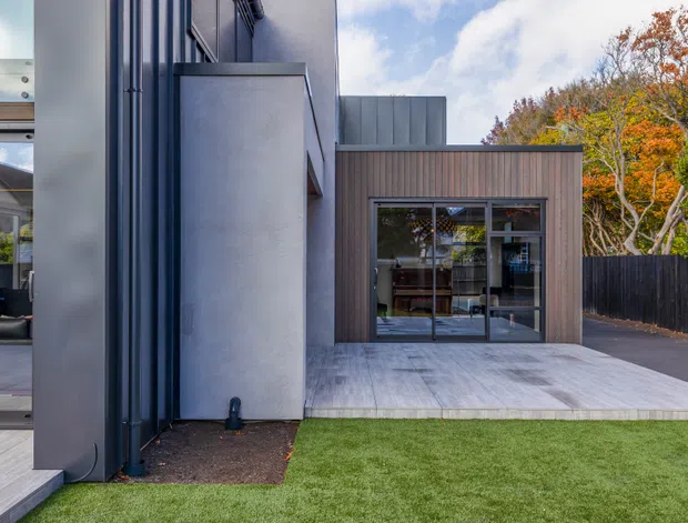 Exterior view of a contemporary residential extension with dark vertical cladding, rendered wall panels, large glazed sliding doors, and a broad timber-look deck opening onto a small lawn. Autumn trees and a dark boundary fence are visible beyond the property.