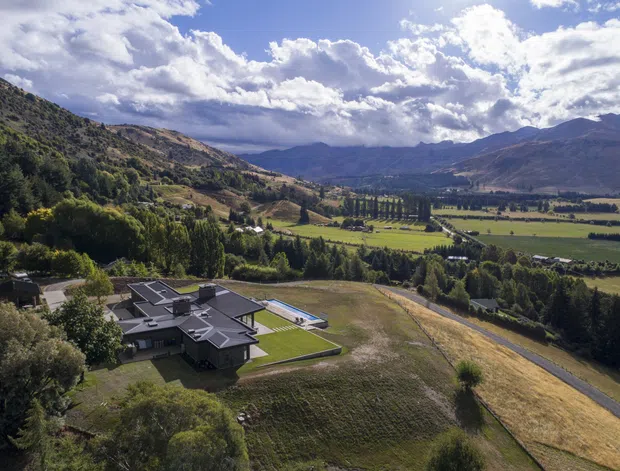 Aerial view of a contemporary low-profile residence set within a spacious rural valley landscape. The dark, angular roof forms and pale exterior walls are visible in the foreground, with manicured lawns, surrounding fields, tree-lined roads, and dramatic mountain ranges beyond.