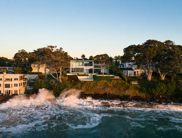 Aerial coastal view of a contemporary residential property positioned directly above a rocky shoreline, with waves breaking against the coast. The main house has a rectilinear form, extensive glazing, pale exterior finishes, and landscaped terraces, with neighbouring homes and mature trees visible along the waterfront.