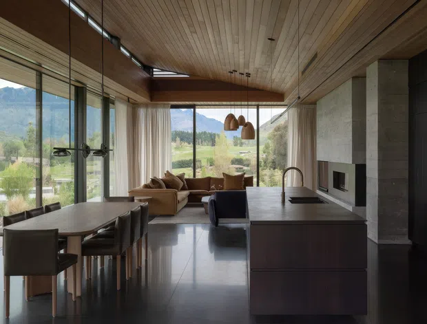 Open-plan contemporary kitchen, dining, and living space with extensive floor-to-ceiling glazing overlooking a mountainous rural landscape. The room features a large dark kitchen island, integrated sink and tap, timber-lined ceiling and walls, polished dark flooring, a concrete fireplace wall, dining table, lounge seating, and pendant lighting. The design has a warm, refined natural-material palette and strong indoor-outdoor visual connection.